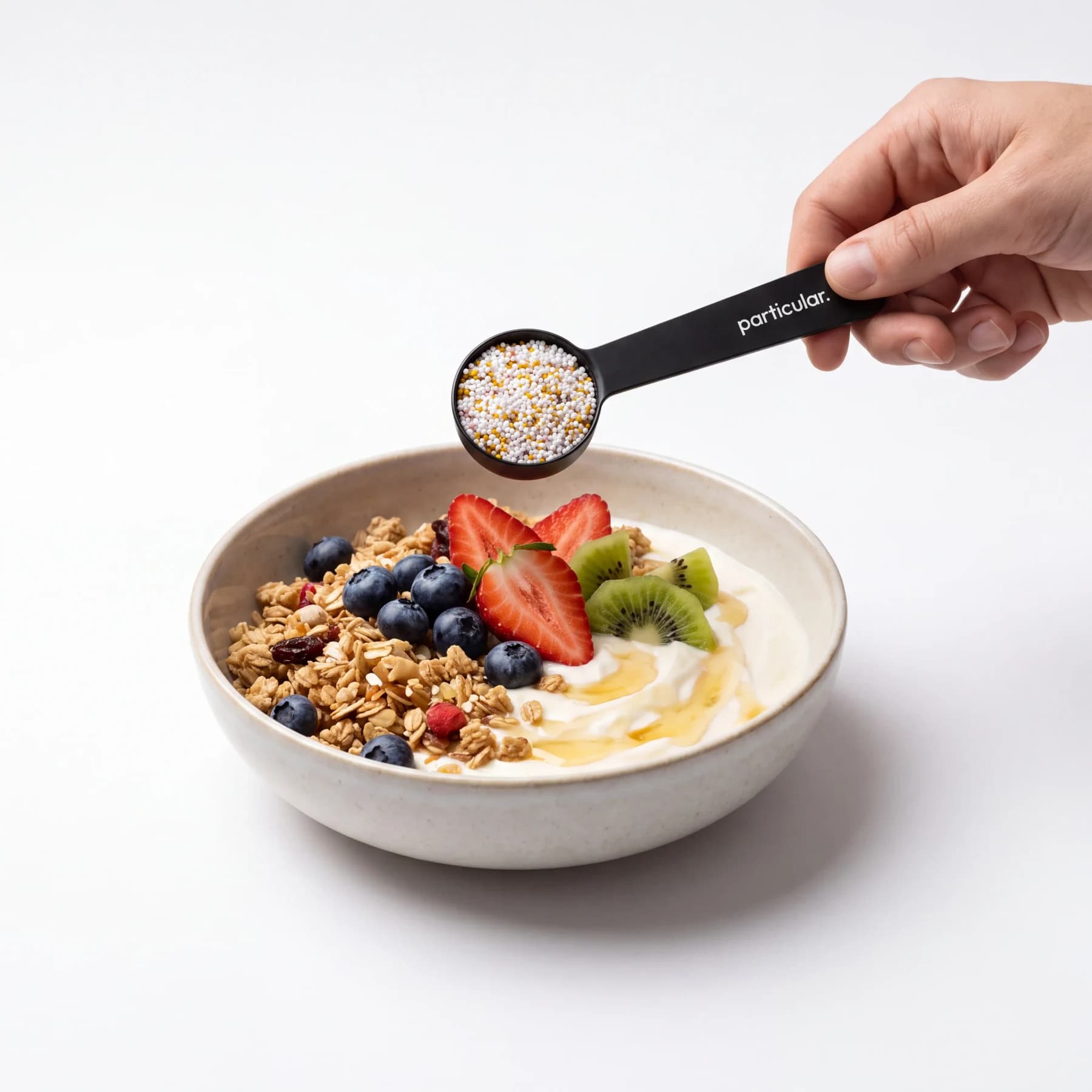 A hand holding a Particular scoop of granules above a bowl of yoghurt, granola and fruit